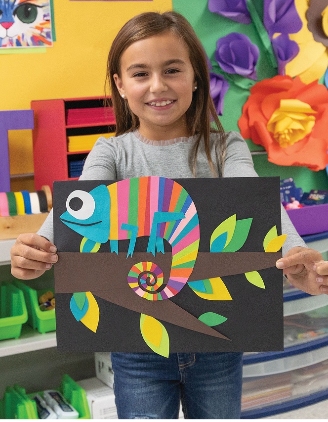 Girl holding colorful chameleon artwork in a bright classroom setting.