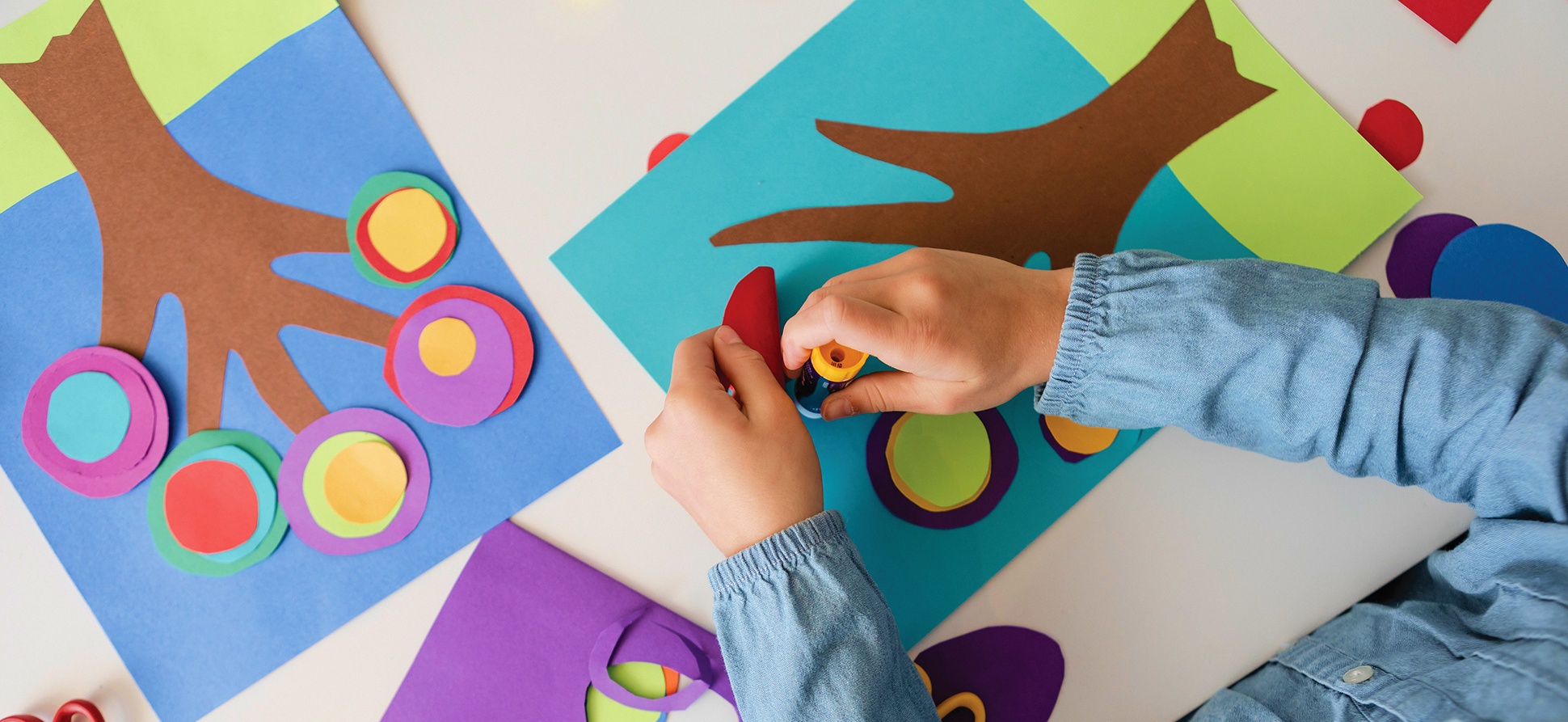 Child creating colorful art with paper shapes and glue on a table.