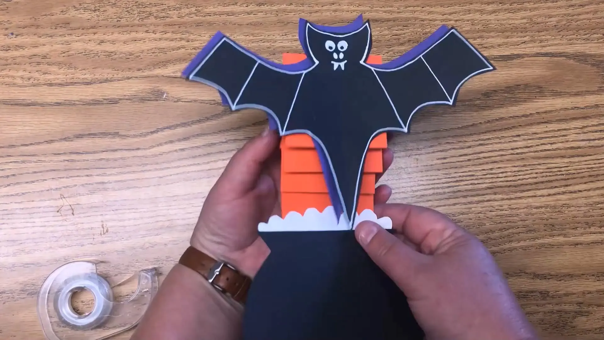 Hands assembling a paper bat craft from black, purple and orange Tru-Ray construction paper